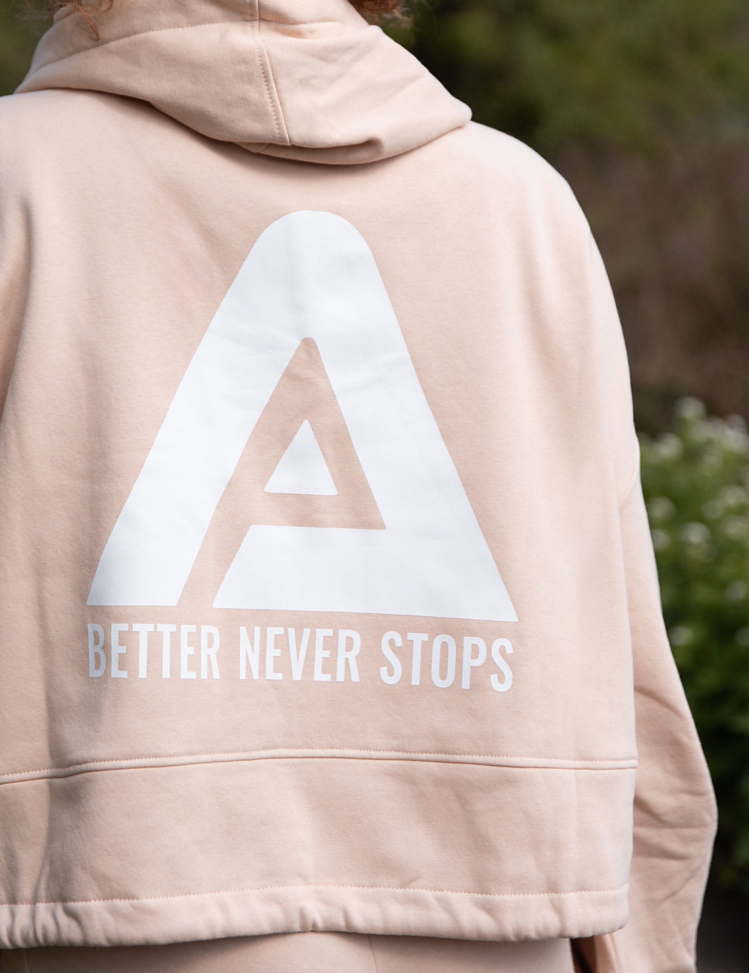 Back view of women's soft pink oversized crop hoodie with tie waist and white Better Never stops logo.